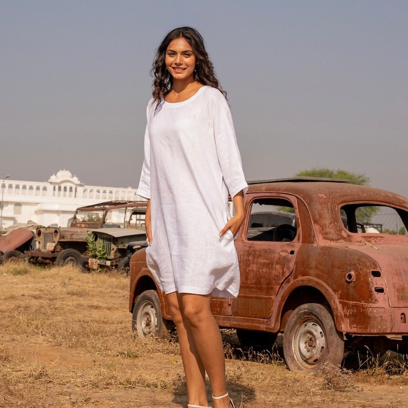 Puede incluir: Una mujer con un vestido blanco de lino est&aacute; de pie frente a un coche oxidado. El vestido tiene un escote redondo y mangas tres cuartos. La mujer sonr&iacute;e y mira a la c&aacute;mara.