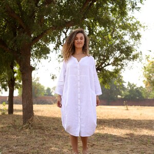 May include: A woman with curly hair is wearing a white linen dress with a button-down front. She is standing in a field with trees in the background.