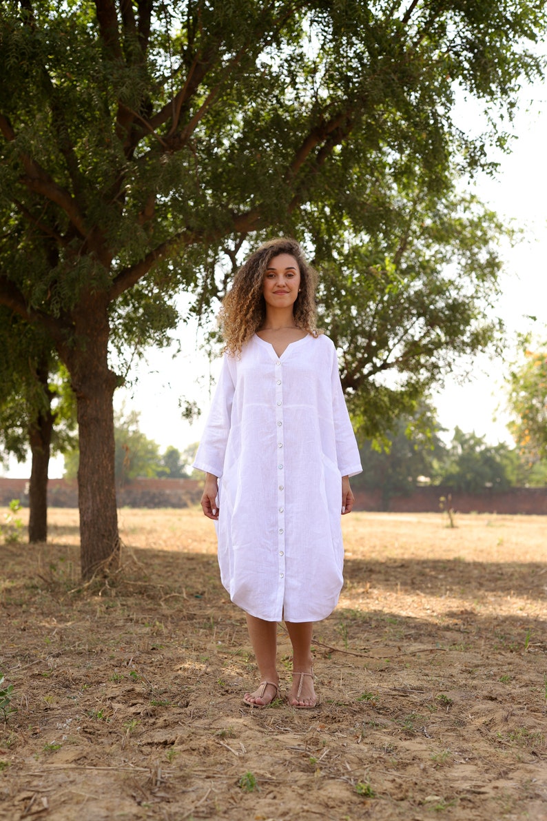 May include: A woman with curly hair is wearing a white linen dress with a button-down front. She is standing in a field with trees in the background.