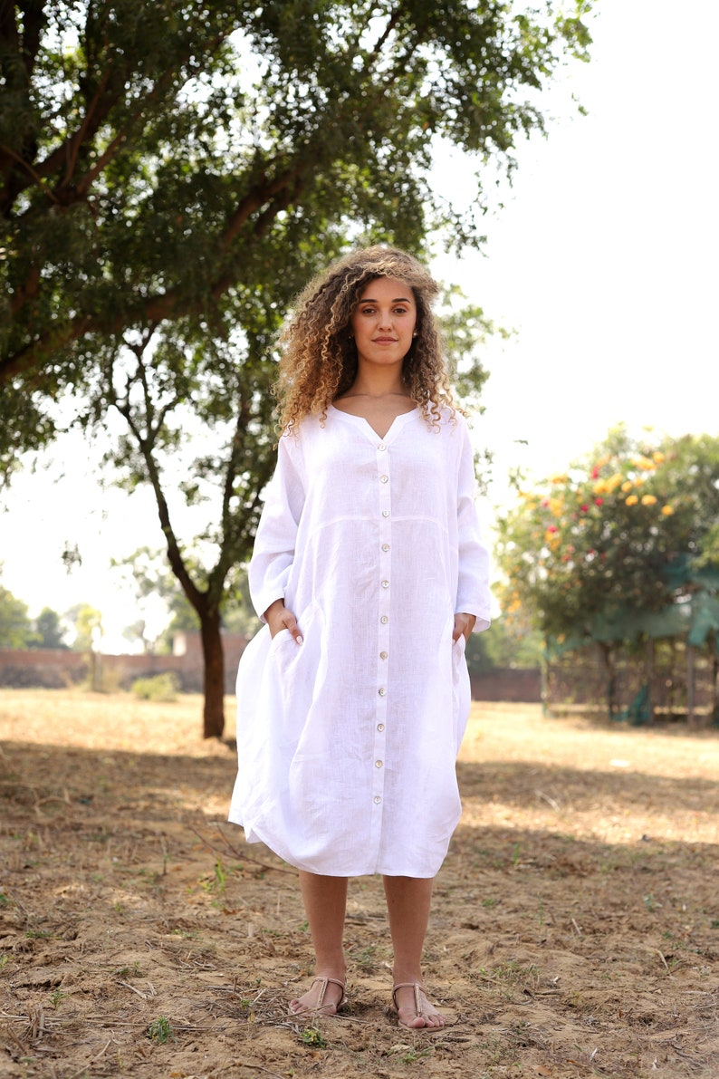 May include: A woman wearing a white linen dress with a button-down front and long sleeves. She is standing in a field with a tree in the background.