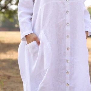 May include: A white linen shirt dress with a button-up front and a large pocket on the left side.