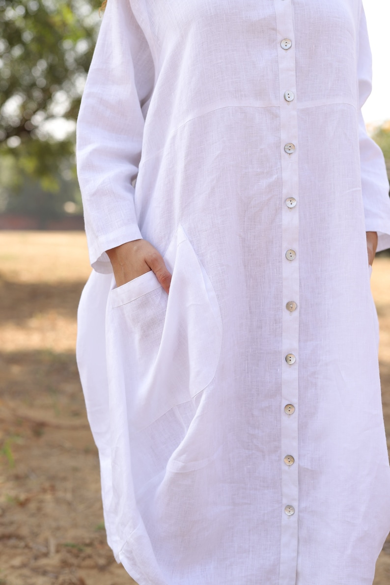 May include: A white linen shirt dress with a button-up front and a large pocket on the left side.
