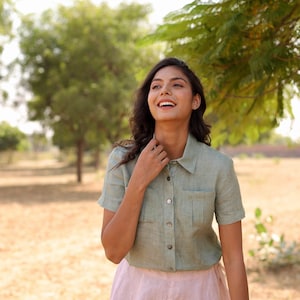 Puede incluir: Una mujer lleva una camisa de lino verde claro con cuello y mangas cortas. La camisa tiene cierre de botones y un bolsillo de parche en el pecho. Ella está sonriendo y mirando hacia arriba.
