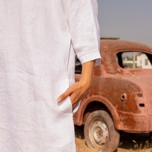 Puede incluir: Una camisa blanca de lino con un bolsillo en el lado izquierdo. La camisa la lleva puesta una persona con la mano en el bolsillo. La persona est&aacute; de pie delante de un coche rojo oxidado.