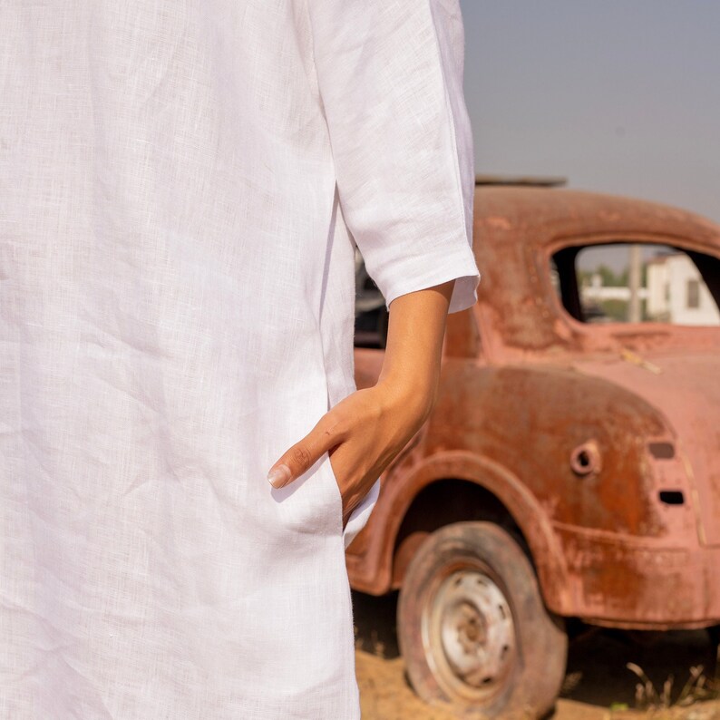 Puede incluir: Una camisa blanca de lino con un bolsillo en el lado izquierdo. La camisa la lleva puesta una persona con la mano en el bolsillo. La persona est&aacute; de pie delante de un coche rojo oxidado.