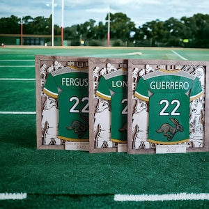 May include: Three framed wooden art pieces featuring green and gold football jerseys with player names and the number 22. Each jersey has a rabbit logo. The text "Quincy Jacks Assistant 2022" is at the bottom of each frame.