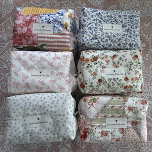 May include: A collection of quilted cosmetic bags in various floral and patchwork patterns. Each bag features a small label with the text "Enchanted Fiber&Co." and a heart symbol. The bags are in shades of blue, red, pink, and cream.