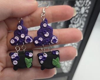 May include: Handmade purple and black polymer clay earrings. The earrings feature a cauldron design with green leaves and a purple cloud-like shape with white and purple accents. The earrings are held by a person.