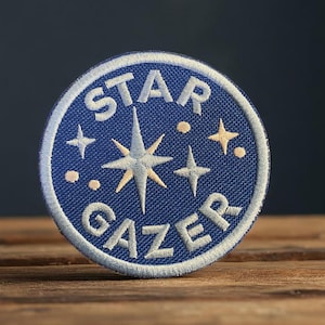 May include: A circular blue embroidered patch with the words "STAR GAZER" in white lettering. The patch features a central starburst design and smaller star and dot accents. The patch is on a wooden surface.