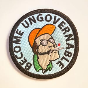 May include: Embroidered patch with a cartoon image of a man wearing an orange hat and sunglasses, smoking a cigarette. The patch has a light blue background and a black border. The words "BECOME UNGOVERNABLE" are embroidered around the image.