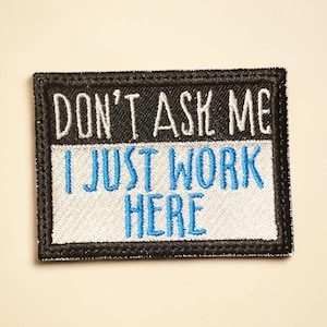 May include: Embroidered patch with the text "DON'T ASK ME I JUST WORK HERE" in white and blue. The patch has a black border and is rectangular in shape. The background is a light beige color.