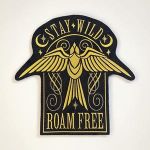 Pode incluir: Patch bordado com fundo preto e design dourado. O patch apresenta um pássaro estilizado com as asas abertas, as palavras "STAY WILD" e "ROAM FREE". O design inclui estrelas e detalhes de lua crescente.