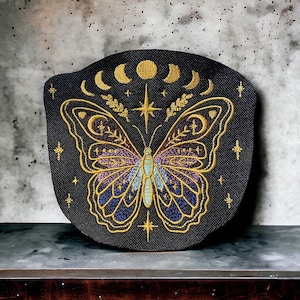 Puede incluir: Un parche bordado negro con un diseño de mariposa dorada. La mariposa tiene alas moradas y azules con un contorno dorado. El parche también presenta una estrella dorada y lunas crecientes.