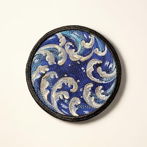 May include: Round, embroidered patch featuring a stylized ocean wave design in shades of blue and white. The patch has a black border and is likely for decorative purposes, such as on clothing or bags.