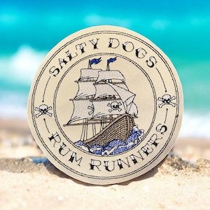 Op de afbeelding: Een ronde, beige patch met geborduurde tekst die "Salty Dogs Rum Runners" luidt. De patch toont een gestileerd schip met zeilen en een schedel met gekruiste beenderen. Het schip vaart over blauwe golven.