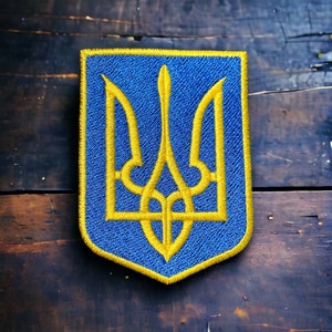 May include: A blue and gold embroidered patch featuring the Ukrainian coat of arms, a trident, on a shield-shaped background.
