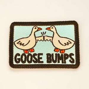 May include: Embroidered patch with two cartoon geese bumping beaks. The geese are cream-coloured with orange beaks and feet, set against a light blue background. The words "GOOSE BUMPS" are in black, below the geese, with a black border.