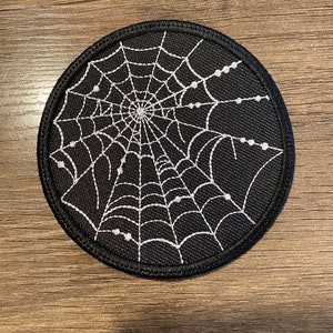 May include: A black round patch with a white embroidered spider web design. The web has a central hub and radiating lines with small white dots at the ends.