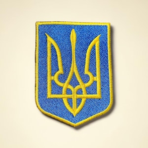 May include: Embroidered patch featuring the Ukrainian coat of arms, a gold trident on a blue shield. The patch has a gold border and is likely for clothing or accessories. The design is a symbol of Ukrainian national identity.