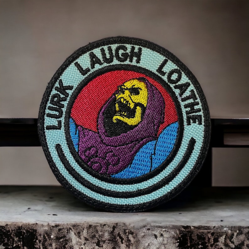 May include: A round, blue and black embroidered patch with the words "Lurk Laugh Loathe" around the edge. In the center is a cartoon image of Skeletor from the Masters of the Universe cartoon, with a purple and blue robe, a yellow skull, and a red background.