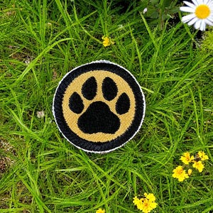 Op de afbeelding: Ronde patch met een zwarte pootafdruk op een gouden achtergrond, omgeven door een zwart-witte rand. De patch ligt op groen gras, met kleine gele bloemen en witte madeliefjes op de achtergrond.