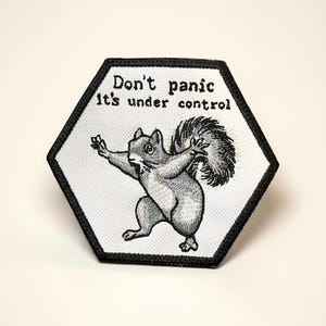 May include: Hexagon-shaped patch with a black border and the text "Don't panic it's under control." The patch features a cartoon squirrel with its arms outstretched. The squirrel is gray and white, with a fluffy tail.