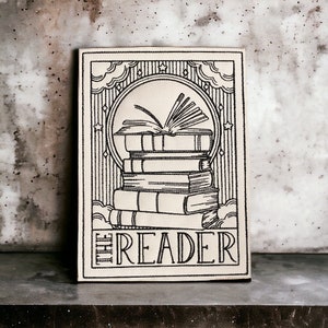 May include: A black and white illustration of a stack of books with an open book on top. The illustration is on a beige background with the text "THE READER" at the bottom.
