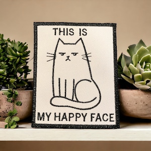 May include: Black and white embroidered patch featuring a cat and the text "This is my happy face".
