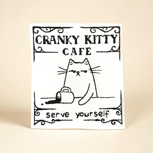 May include: A white square patch with a black outline featuring a grumpy cat illustration. The text "CRANKY KITTY CAFE" and "serve yourself" are displayed in black. A spilled coffee cup is also shown.