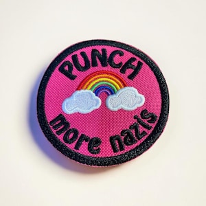 May include: A circular, pink embroidered patch with the words "PUNCH more nazis" in black, curved around the top and bottom. A rainbow and two white clouds are in the centre. The patch has a black border.