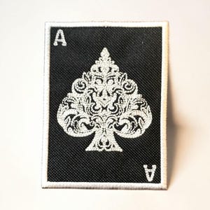 May include: Black and white Ace of Spades patch. The patch features a detailed, ornate spade design in white against a black background. The letter "A" is in the top left and bottom right corners. The patch has a white border.