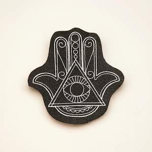 Könnte beinhalten: Schwarzer Hamsa-Hand-Aufnäher mit weißen Stickdetails. Das Design beinhaltet ein Auge in einem Dreieck und dekorative Linien. Der Aufnäher ist ein Symbol für Schutz und Glück.