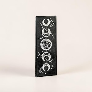 May include: Black rectangular patch featuring a white embroidered design of the phases of the moon and constellations. The patch is a vertical rectangle with a black background and white stitching. The design includes crescent moons and circular shapes.
