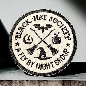 Könnte beinhalten: Schwarz-weiß gestickter Aufnäher mit dem Text "Black Hat Society A Fly By Night Group" und einem Hexenhut, gekreuzten Besen, einer Mondsichel und einem Kessel.