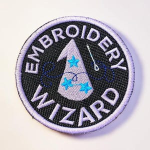 May include: Embroidered patch with the words "EMBROIDERY WIZARD" in white, encircling a design of a wizard hat with blue stars and a needle and thread. The patch is round, with a black center and a purple border.