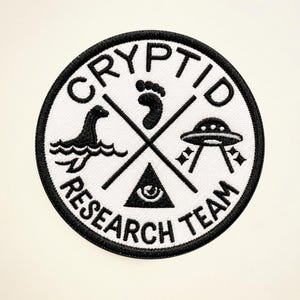 May include: A circular patch with a black border and white centre, embroidered with the words 'CRYPTID RESEARCH TEAM'. The design includes a Loch Ness Monster, a footprint, a UFO, and an eye within a triangle, all in black.