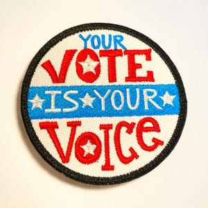 Può includere: Toppa ricamata circolare con la scritta "YOUR VOTE IS YOUR VOICE" in rosso e blu. La toppa ha uno sfondo bianco, un bordo nero e dettagli a stella.