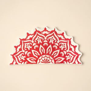 May include: A half-circle, embroidered decorative piece featuring a red mandala design on a white background. The intricate floral pattern is symmetrical and detailed, with a scalloped edge. The piece is set against a cream-colored backdrop.