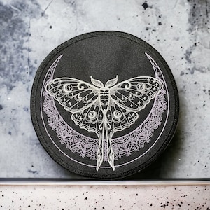 May include: A black fabric patch with a white embroidered moon and moth design. The moth has a detailed design with crescent moons and stars. The moon is surrounded by a delicate lace pattern.