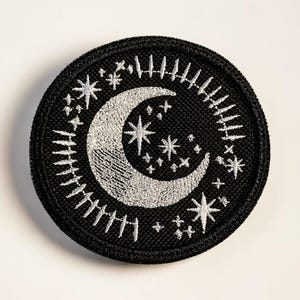 May include: A round, black embroidered patch featuring a crescent moon and stars in silver thread. The design includes a crescent moon, several stars, and a border of vertical lines, creating a celestial theme.