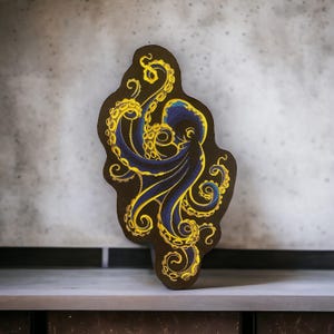 May include: A blue and yellow embroidered octopus patch. The octopus is curled up with its tentacles reaching out. The patch is on a brown background.