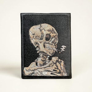 May include: Black wallet with an embroidered image of a skull smoking a cigarette. The skull is beige and tan, set against a black background. The wallet has a rectangular shape.