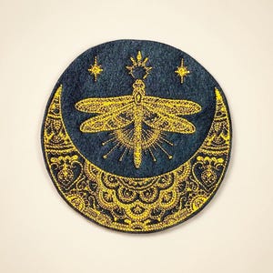 May include: A circular, navy blue patch with a gold embroidered design. The design features a dragonfly in the center, a crescent moon at the bottom, and three stars. The dragonfly has detailed wings and body.