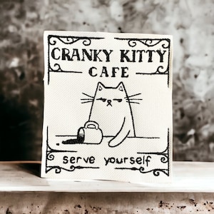 Op de afbeelding: Een wit bord met zwarte tekst en een cartoonkat. Het bord staat er "Cranky Kitty Cafe" en "Bedien uzelf".