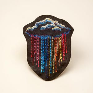May include: A black, shield-shaped patch featuring an embroidered cloud with colorful rain. The cloud is detailed with blue, white, and gold thread, while the rain is depicted in vibrant red, blue, yellow, and orange strands.