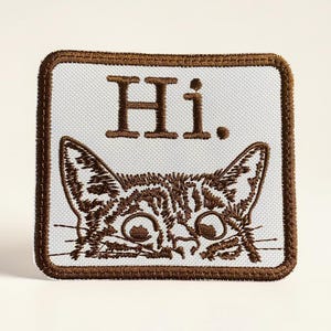 May include: Embroidered patch featuring a cat's face peeking from the bottom. The patch has a white background with the word "Hi." in brown letters at the top. The patch is rectangular with a brown border.