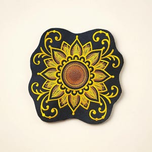 May include: Embroidered patch featuring a detailed sunflower design. The sunflower has yellow petals and a brown center, set against a dark blue, scalloped background. Decorative yellow swirls surround the flower.