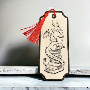 May include: A beige bookmark with a black embroidered illustration of a dragon reading a book while sitting on a stack of books. The bookmark has a red tassel.