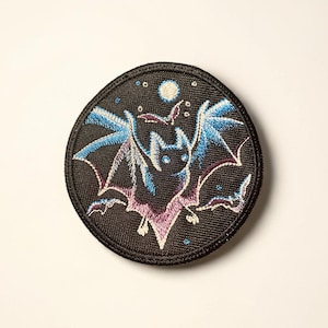 Puede incluir: Parche bordado circular que representa un murciélago con alas azules y moradas sobre un fondo negro con una luna y estrellas. El murciélago es el foco central, con costuras detalladas.