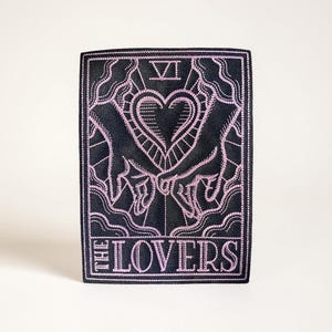 May include: A black and pink embroidered patch depicting the Lovers tarot card. The design features two hands holding a heart, with the words "THE LOVERS" at the base. The patch has a decorative border.